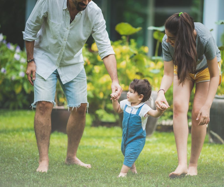 Parents teach toddler to walk in stable environment of back yard. 