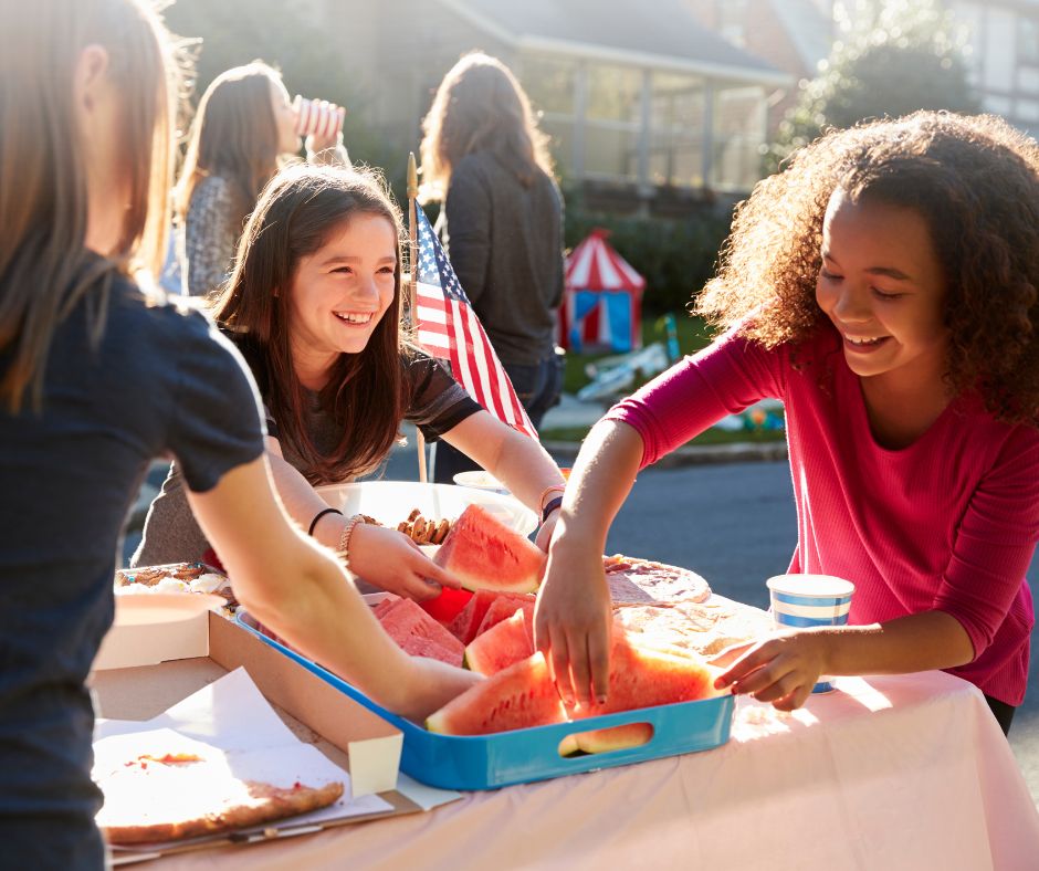 Children bond at a block party, three children over a table of watermelon. 