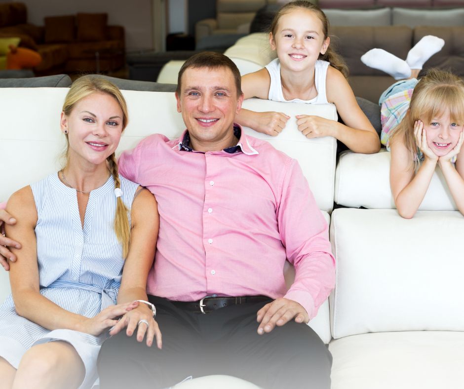 Proud family sitting on sofa in new home, children smile and husbands proud arm wraps around wife.